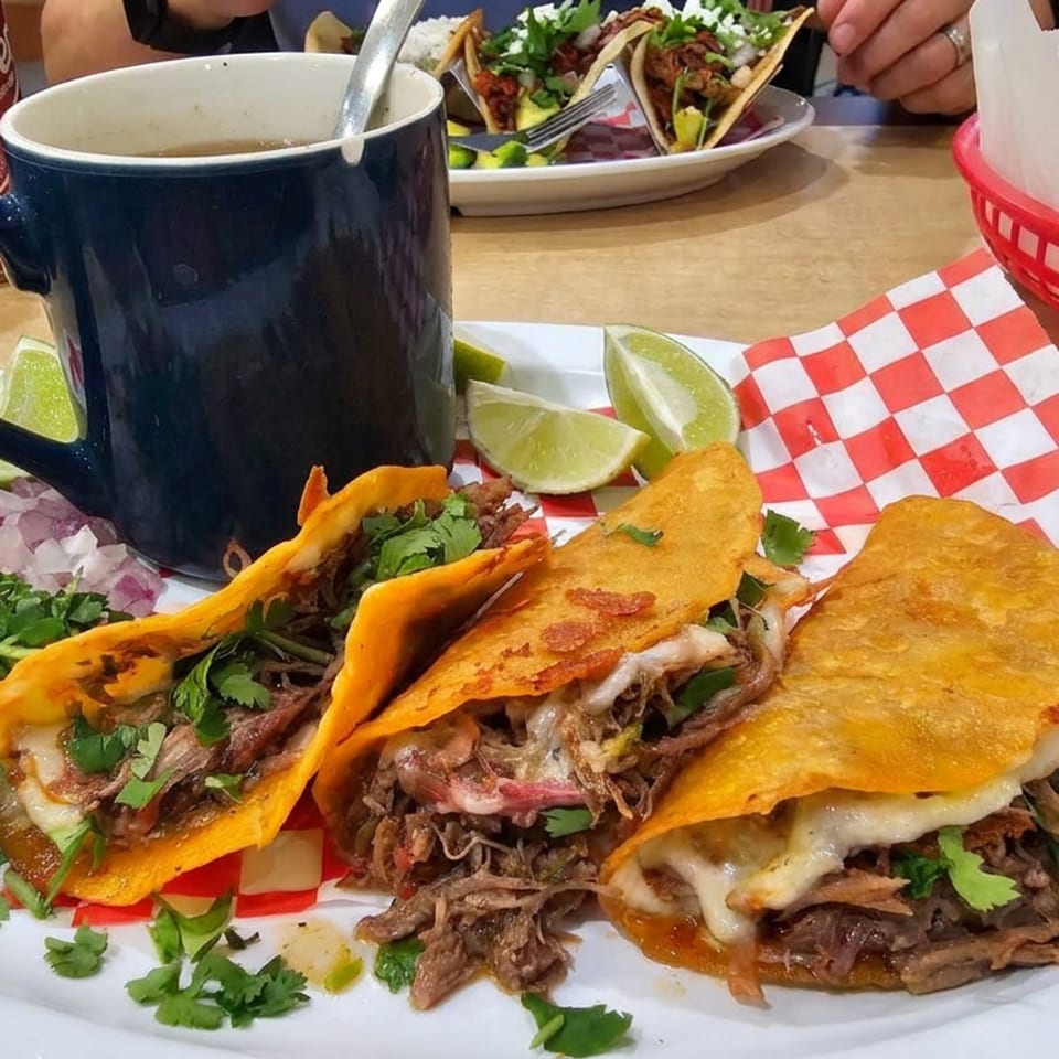 Plate of cheesy shredded-beef tacos with cilantro, onions, lime wedges, and a dark mug on a restaurant table.