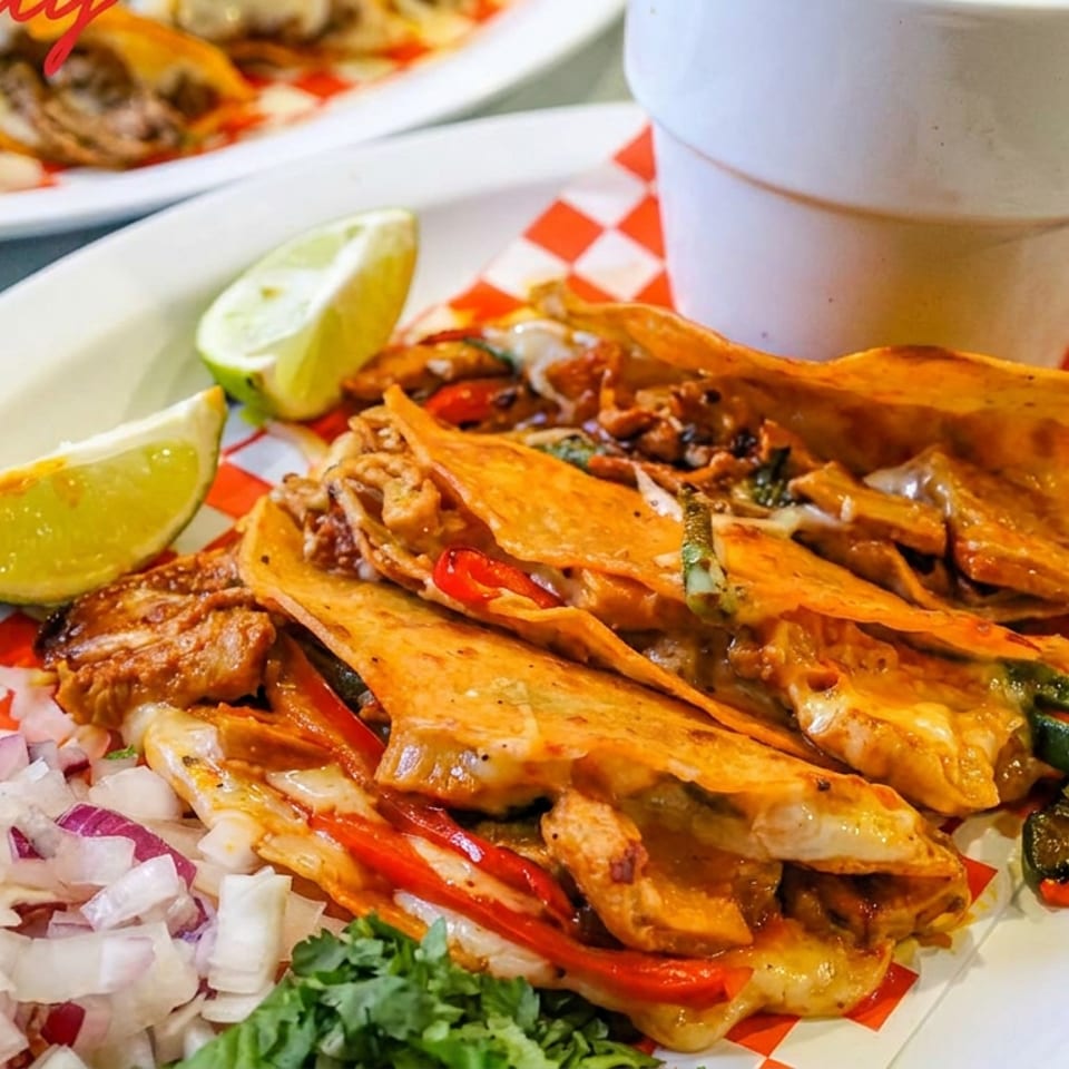 Close-up of cheesy folded tacos with grilled meat, peppers, onions, lime wedges, and cilantro on a checkered-lined plate.
