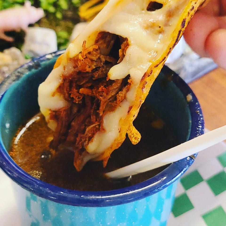 Hand holds a cheesy birria taco over a blue bowl of consommé on a green-and-white checkered table.