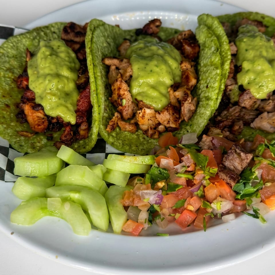 Three green tacos filled with grilled meat and topped with avocado sauce, served with cucumber slices and fresh pico de gallo.